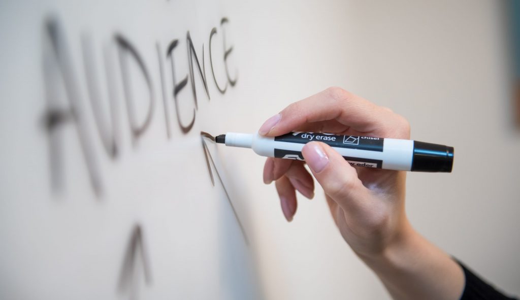 Woman's hand writing the word "audience" on a whiteboard, with arrows.
